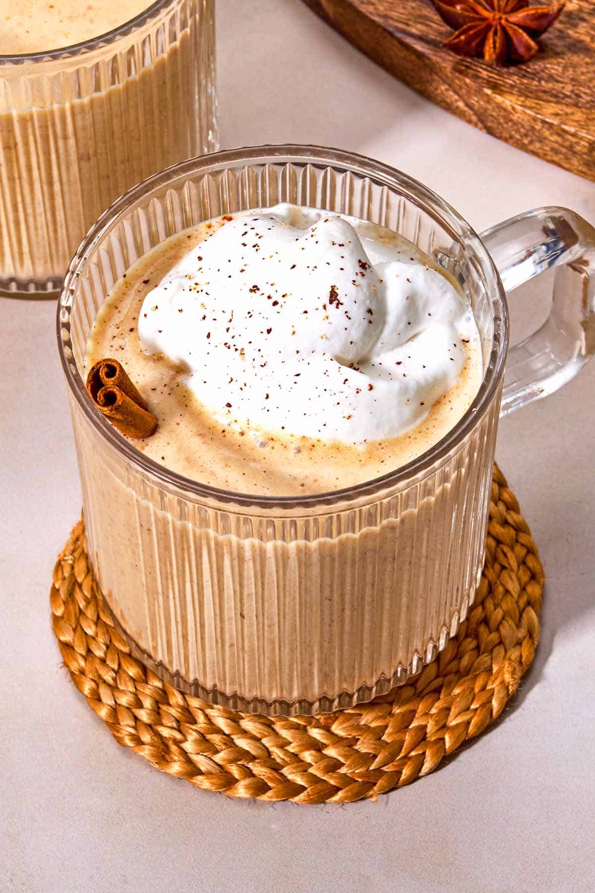 A mug of dairy-free eggnog garnished with a cinnamon stick and whipped coconut cream on a coaster.