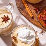 Two mugs of dairy-free eggnog garnished with star anise, a cinnamon stick and whipped coconut cream next to a wooden tray with a bowl of dates, a cinnamon stick, a star anise and the rest of the eggnog in a pitcher.