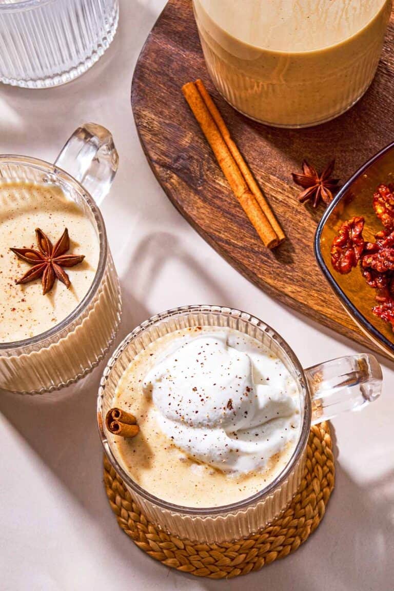 Two mugs of dairy-free eggnog garnished with star anise, a cinnamon stick and whipped coconut cream next to a wooden tray with a bowl of dates, a cinnamon stick, a star anise and the rest of the eggnog in a pitcher.