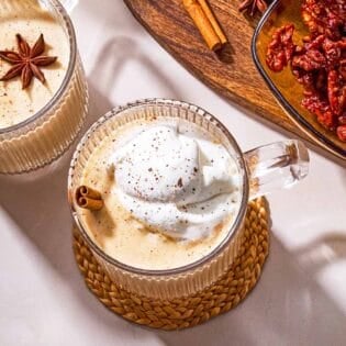 Two mugs of dairy-free eggnog garnished with star anise, a cinnamon stick and whipped coconut cream next to a wooden tray with a bowl of dates, a cinnamon stick a star anise.