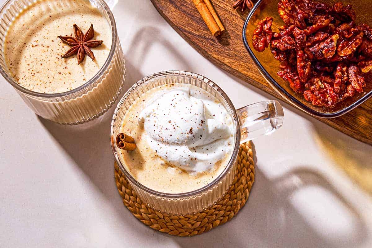 Two mugs of dairy-free eggnog garnished with star anise, a cinnamon stick and whipped coconut cream next to a wooden tray with a bowl of dates, a cinnamon stick a star anise.
