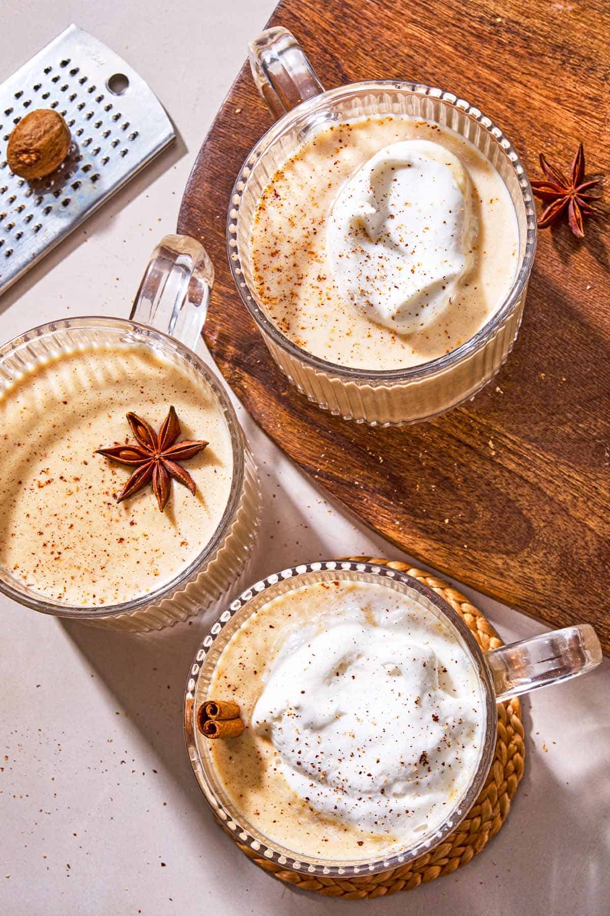Three mugs of vegan eggnog garnished with star anise, a cinnamon stick and whipped coconut cream, one is on a wooden tray. Next to this is a grater with a nutmeg.