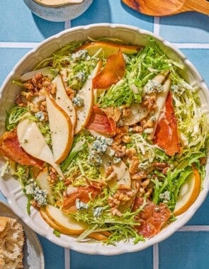 Frisee salad with pears and blue cheese in a serving bowl. Next to this is a plate with crusty bread, a bowl with salt and pepper, a small pitcher of dijon vinaigrette and wooden serving utensils.