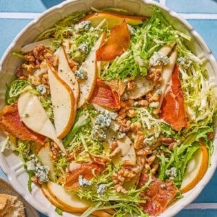 Frisee salad with pears and blue cheese in a serving bowl. Next to this is a plate with crusty bread, a bowl with salt and pepper, a small pitcher of dijon vinaigrette and wooden serving utensils.