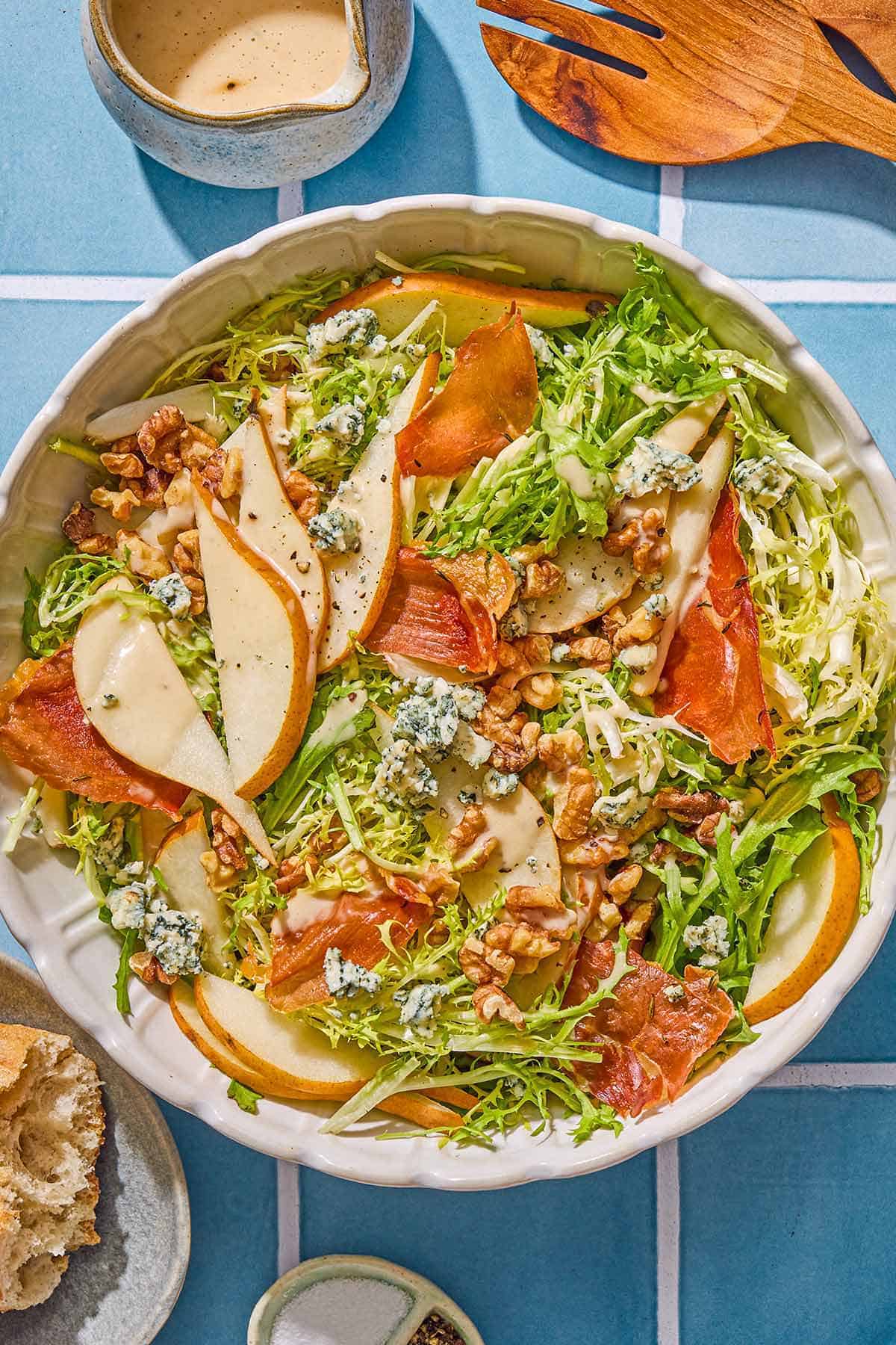 Frisee salad with pears and blue cheese in a serving bowl. Next to this is a plate with crusty bread, a bowl with salt and pepper, a small pitcher of dijon vinaigrette and wooden serving utensils.