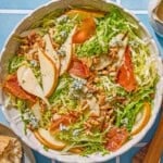 Frisee salad with pears and blue cheese in a serving bowl. Next to this is a plate with crusty bread, a bowl with salt and pepper, a small pitcher of dijon vinaigrette and wooden serving utensils.