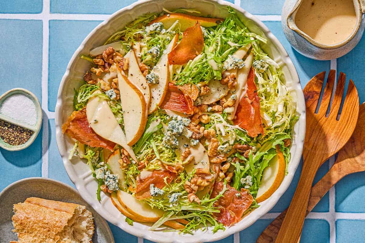 Frisee salad with pears and blue cheese in a serving bowl. Next to this is a plate with crusty bread, a bowl with salt and pepper, a small pitcher of dijon vinaigrette and wooden serving utensils.