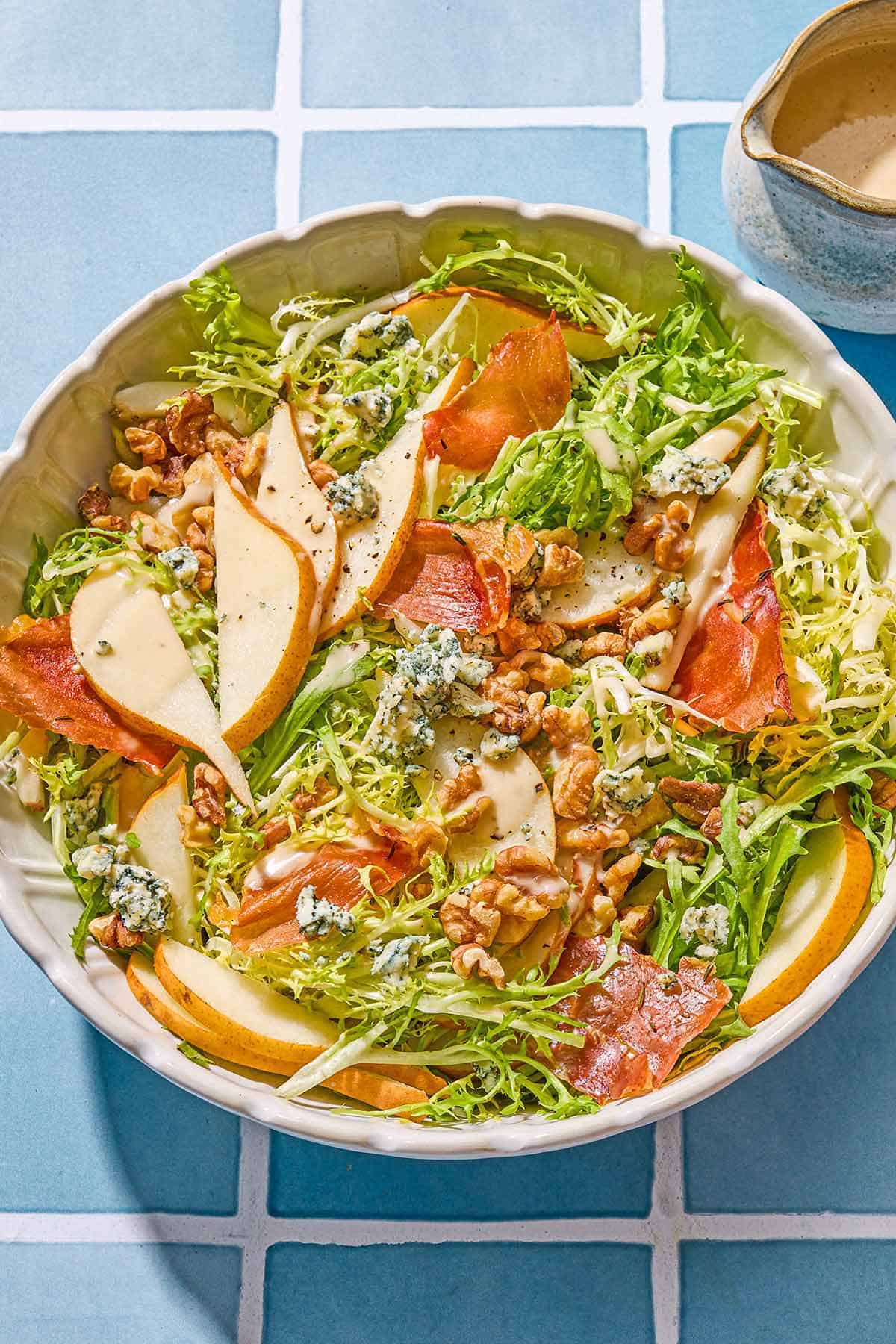 Frisee salad with pears and blue cheese in a serving bowl next to a small pitcher of the dijon vinaigrette.