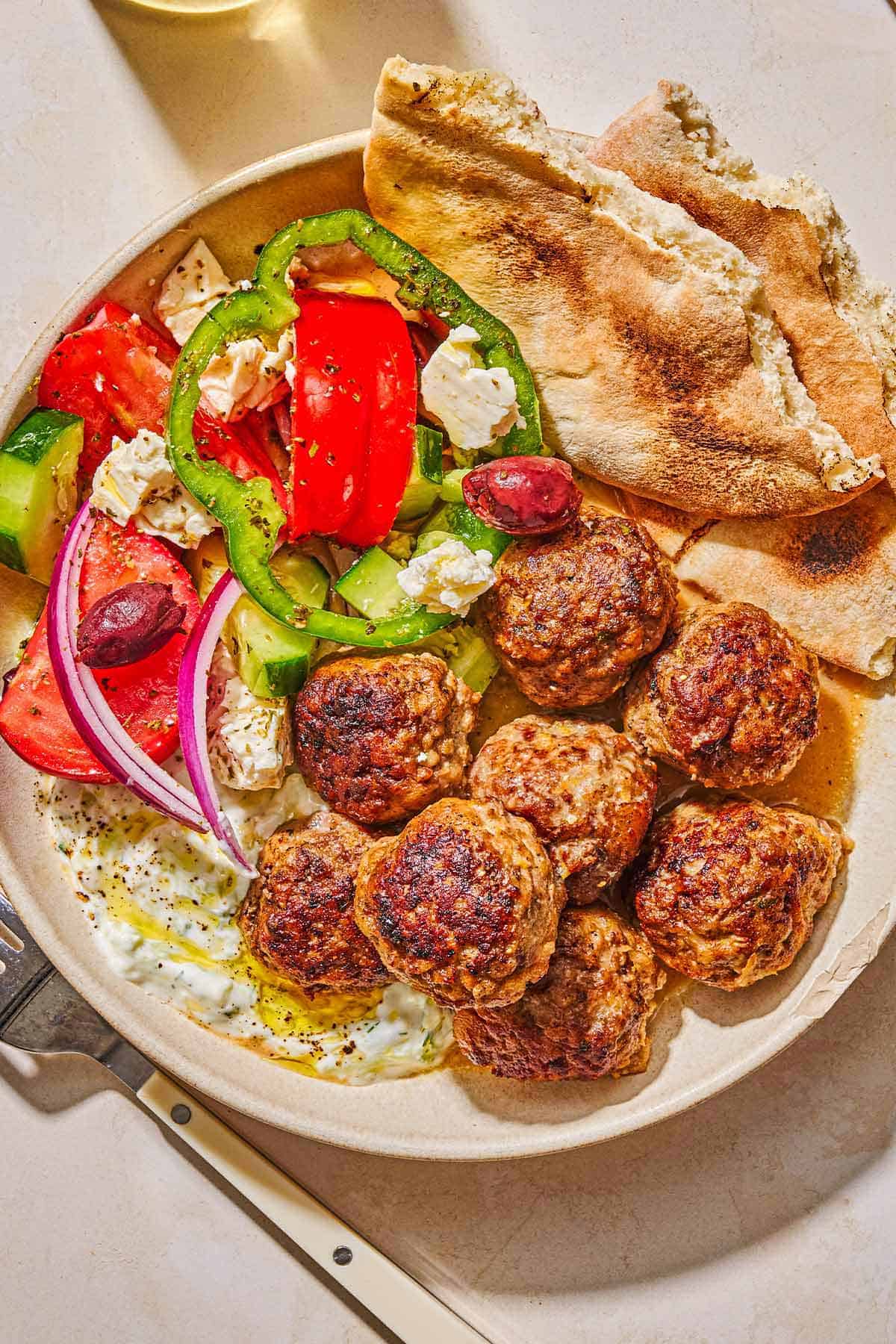 A serving of greek meatballs on a plate with pita, tzatziki sauce and a Greek salad next to a fork.
