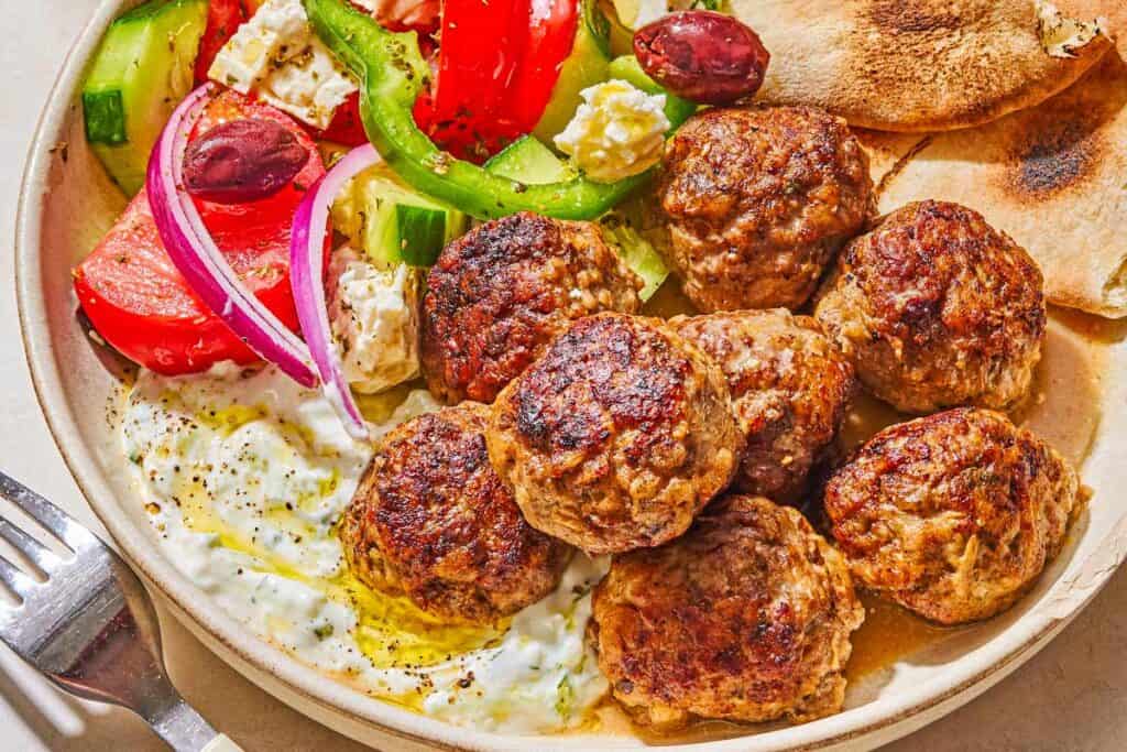 A serving of greek meatballs on a plate with pita, tzatziki sauce and a Greek salad next to a fork.