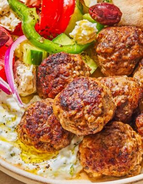 A serving of greek meatballs on a plate with pita, tzatziki sauce and a Greek salad next to a fork.