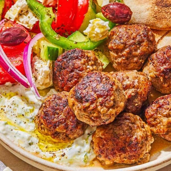 A serving of greek meatballs on a plate with pita, tzatziki sauce and a Greek salad next to a fork.