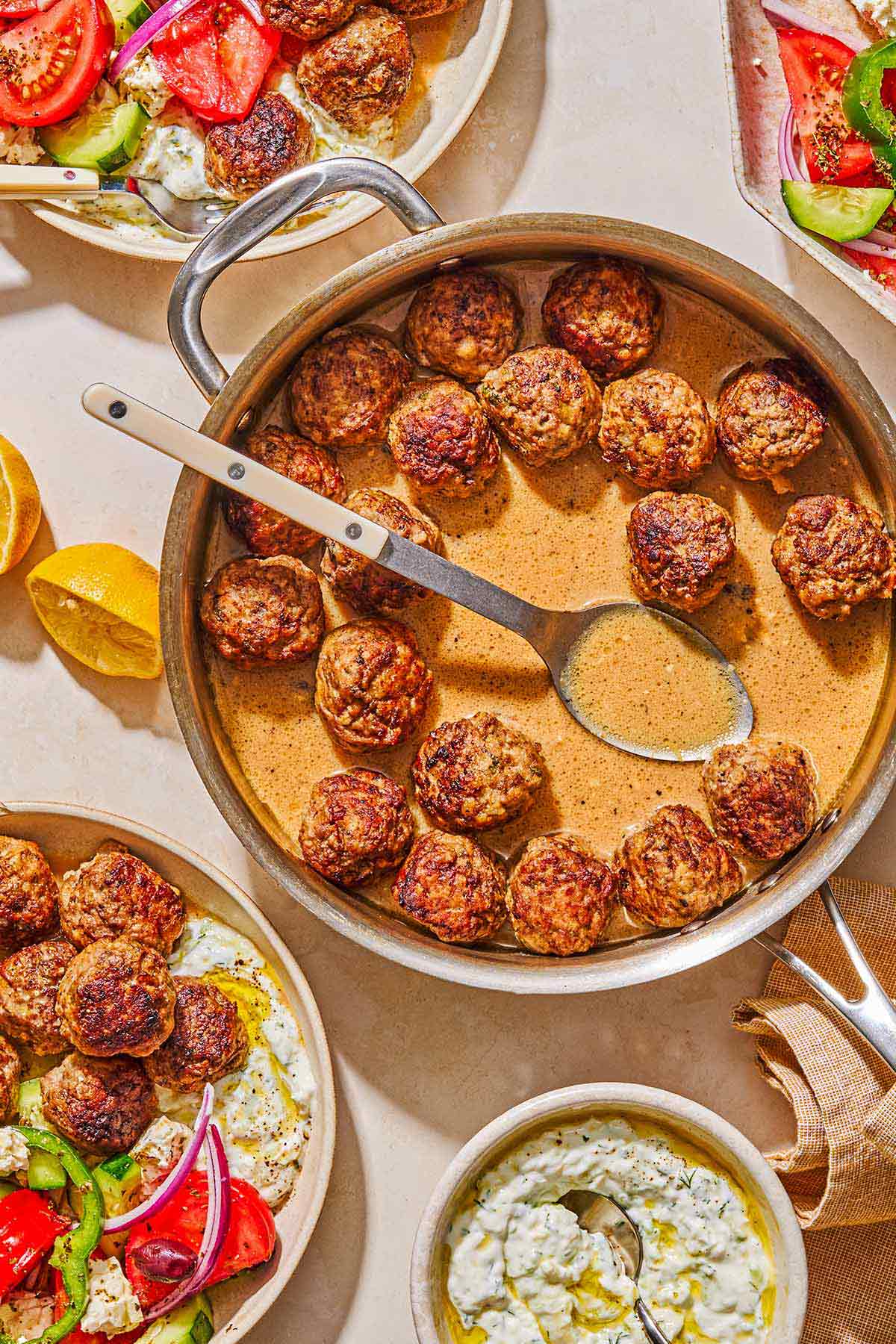 Greek meatballs in a skillet with a serving spoon. Next to this is a bowl of tzatziki sauce, a greek salad, juiced lemon halves, a glass of water, pita bread, and two plates with servings of the greek meatballs, greek salad, tzatziki and pita bread.