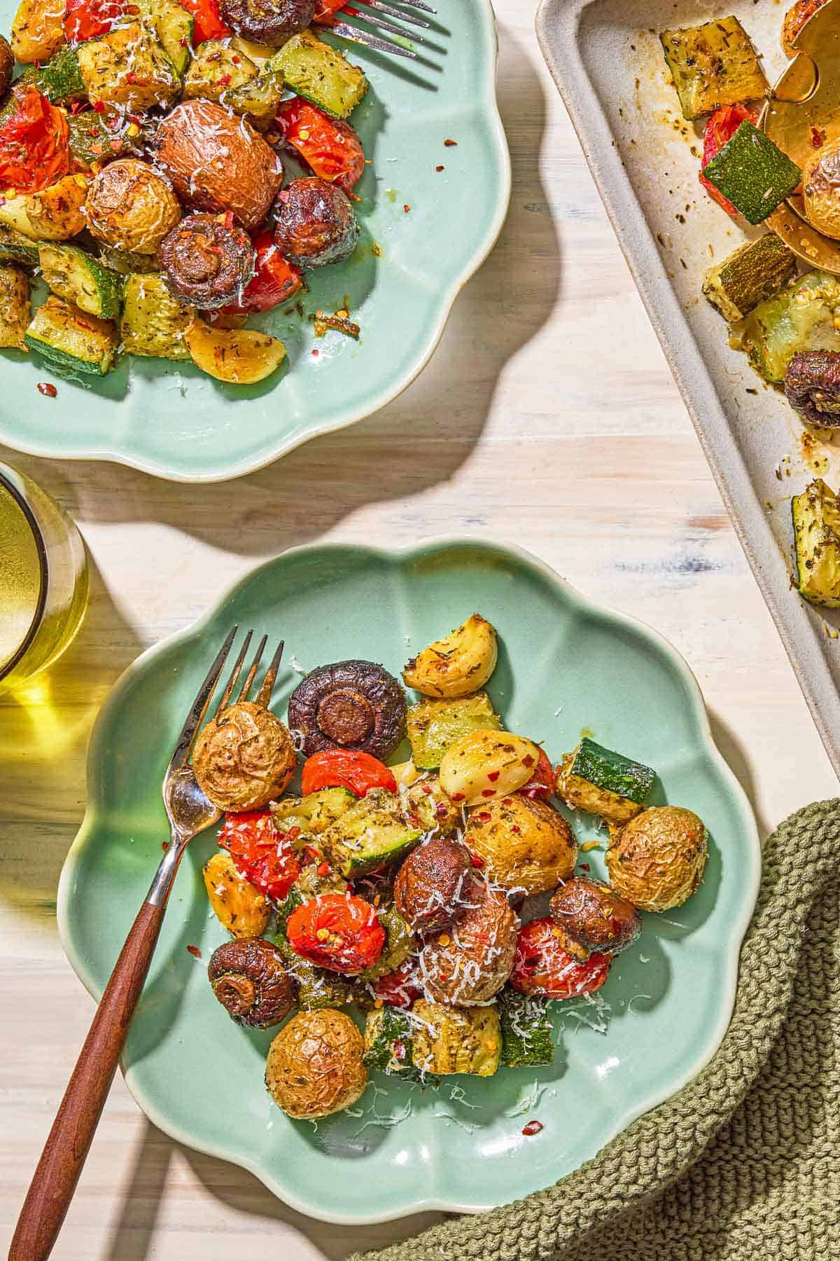 Two servings of the roasted vegetables recipe on plates with forks. Next to these is a kitchen towel, a glass of water and a sheet pan with the rest of the vegetables.