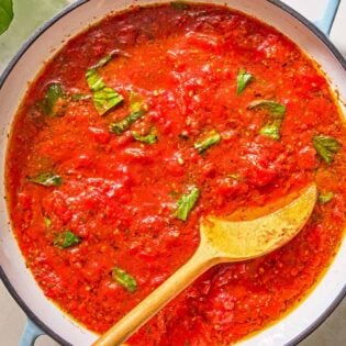 Marinara sauce in a skillet with a wooden spoon. Next to this is a bottle of olive oil and sprigs of fresh basil.
