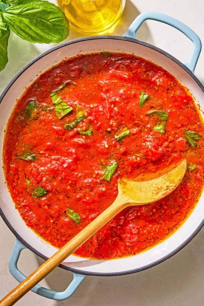 Marinara sauce in a skillet with a wooden spoon. Next to this is a bottle of olive oil and sprigs of fresh basil.