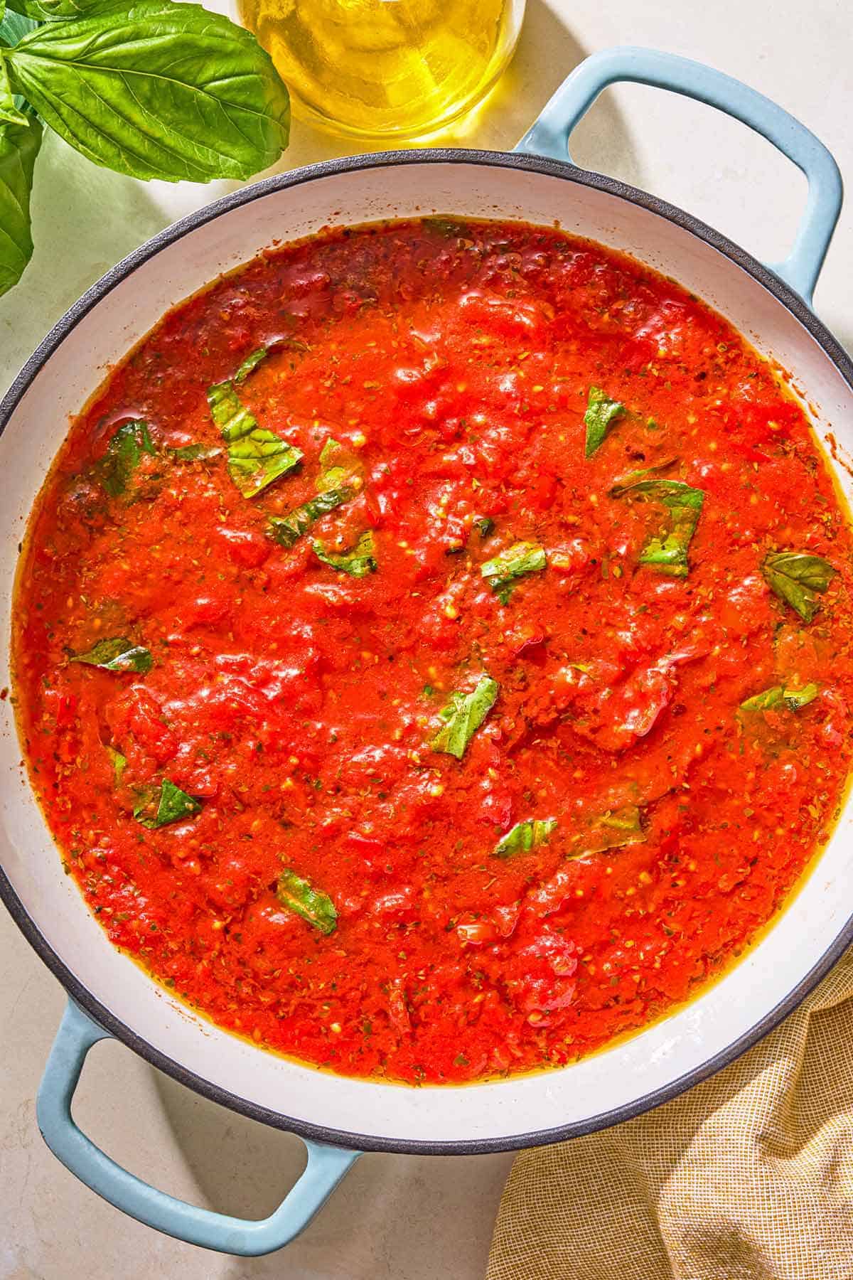 Marinara sauce in a skillet. Next to this is a bottle of olive oil and sprigs of fresh basil.
