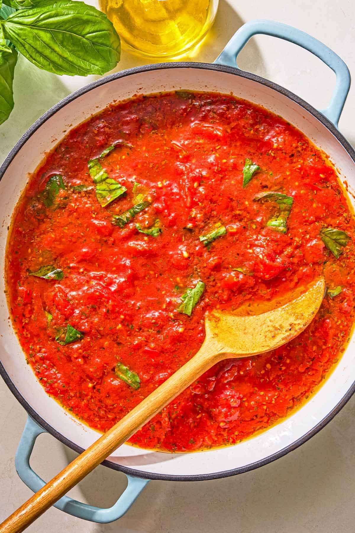 Marinara sauce in a skillet with a wooden spoon. Next to this is a bottle of olive oil and sprigs of fresh basil.