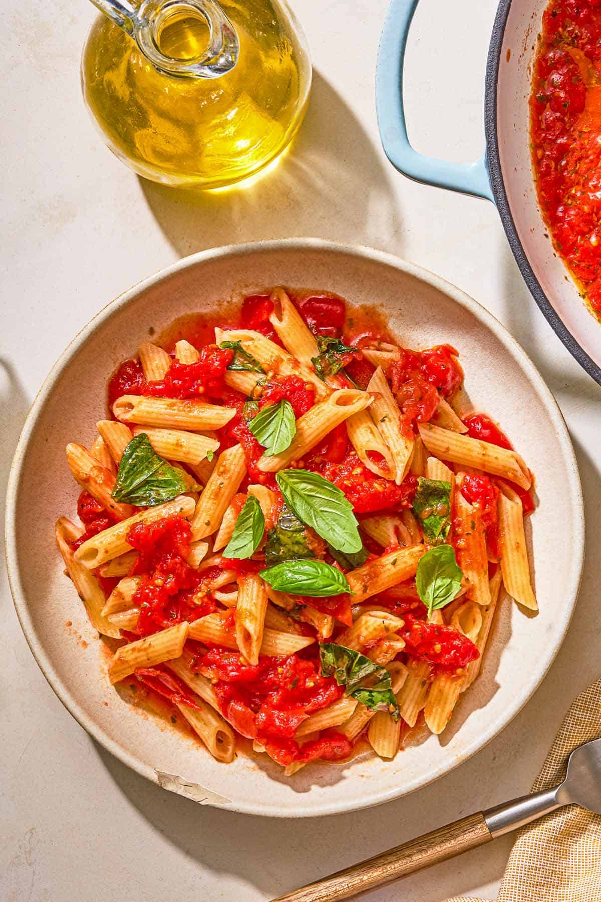 A bowl of pasta tossed in marinara sauce next to a fork, a bottle of olive oil and the rest of the sauce in a skillet.