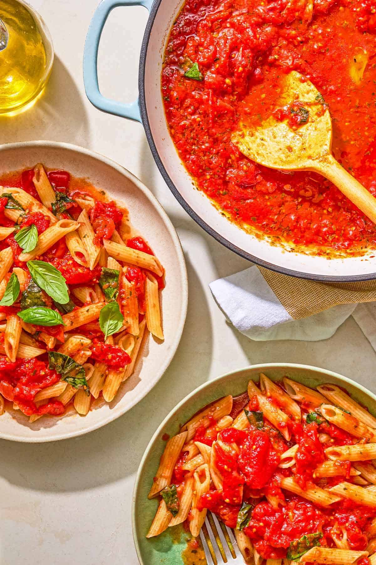 Two bowls of pasta tossed in marinara sauce in bowls. Next to these is the rest of the sauce in a skillet with a wooden spoon and a bottle of olive oil.