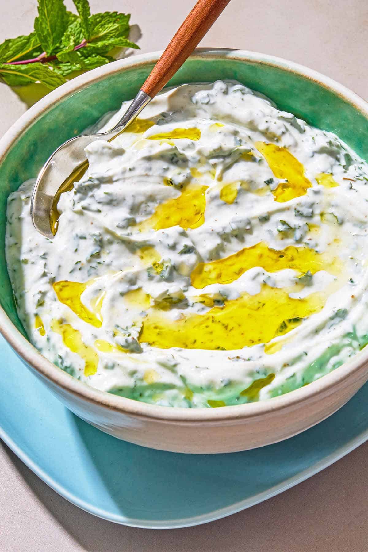 Mint yogurt sauce in a bowl with a spoon on a plate. Next to this is a sprig of mint.