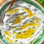 Mint yogurt sauce in a bowl with a spoon. Next to this is a sprig of mint and 2 cloves of garlic.