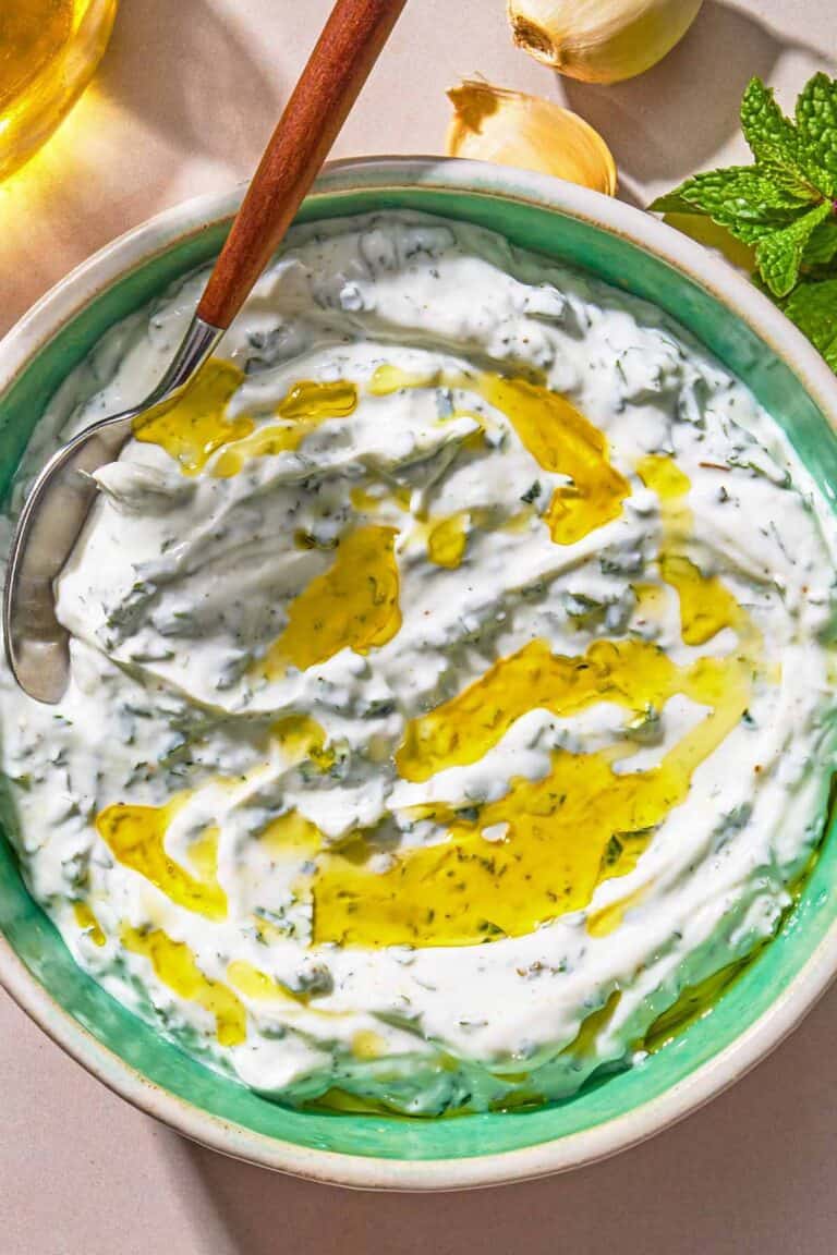 Mint yogurt sauce in a bowl with a spoon. Next to this is a sprig of mint and 2 cloves of garlic.