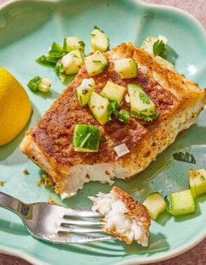 A serving of fried red snapper topped with cucumber salsa on a plate with a fork and a lemon wedge.