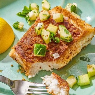 A serving of fried red snapper topped with cucumber salsa on a plate with a fork and a lemon wedge.