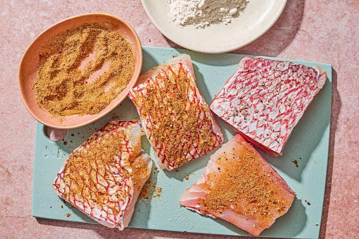 Four snapper fillets on a cutting board with a bowl of the seasoning. The seasoning has been added to 3 of the fillets.