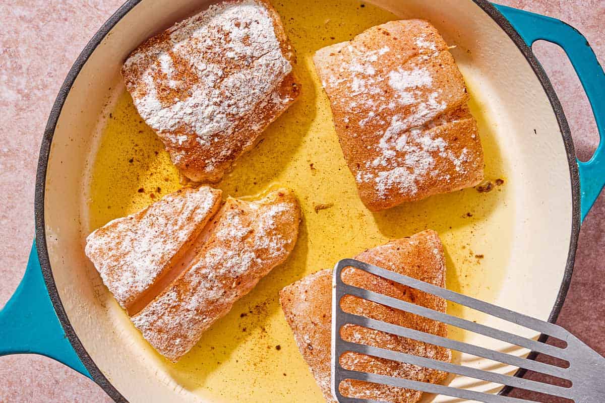 Four red snapper fillets being fried in a skillet with a spatula.