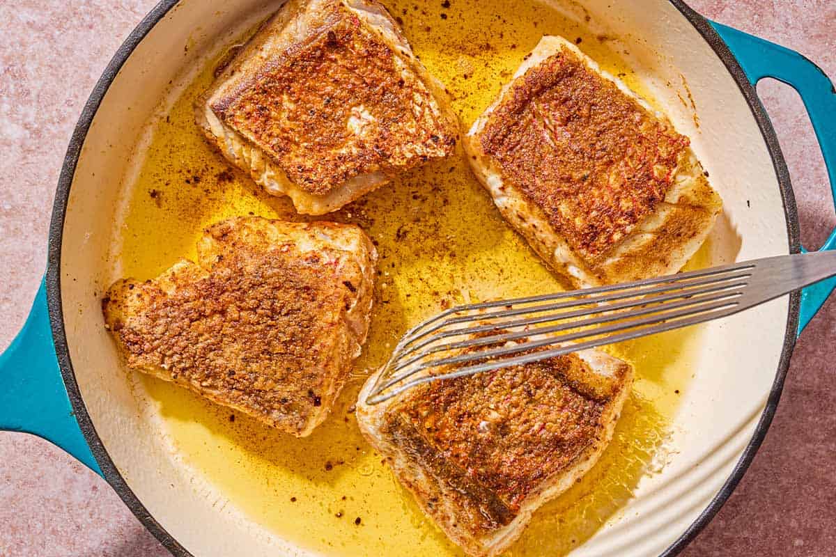 Four red snapper fillets being fried in a skillet with a spatula.