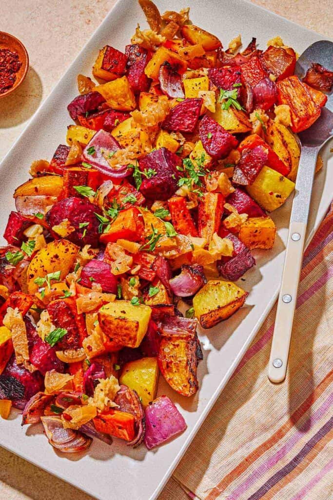 Roasted root vegetables on a platter with a serving spoon. Next to this is a bowl of aleppo pepper.