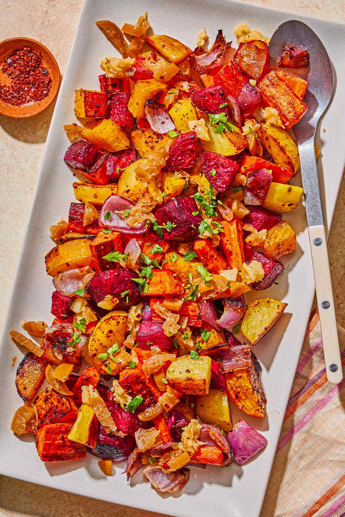 Roasted root vegetables on a platter with a serving spoon. Next to this is a bowl of aleppo pepper.