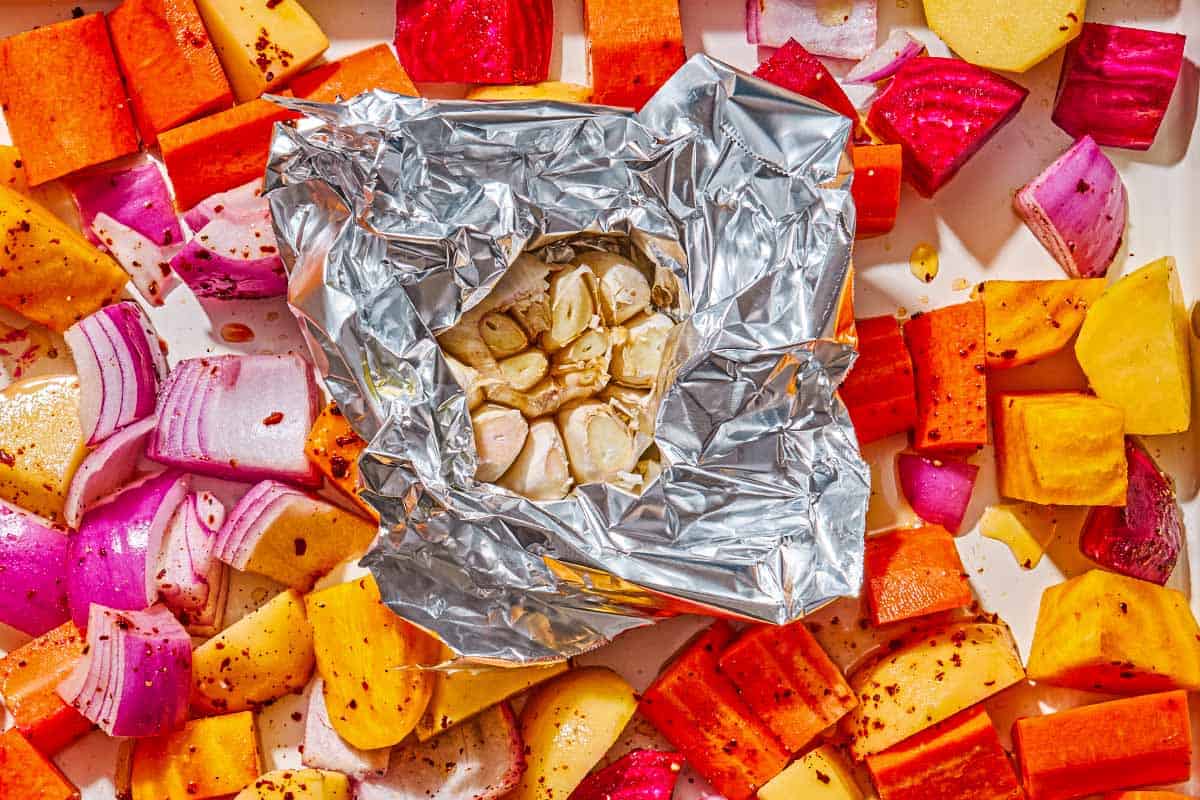 A head of garlic in a piece of foil surrounded by root vegetables on a sheet pan.
