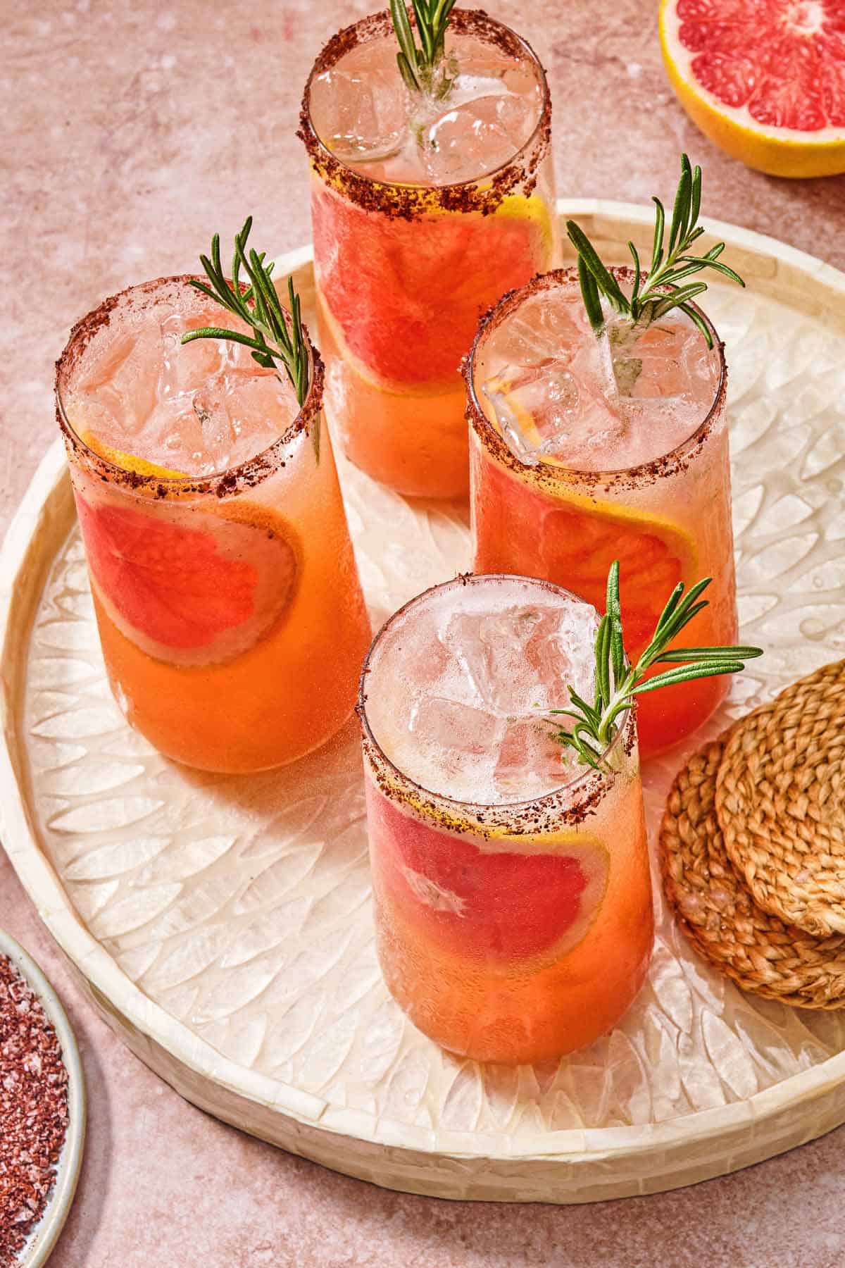 Four glasses of the rosemary grapefruit tonic with grapefruit wheels, sprigs of rosemary and sumac rims on a tray with a stack of coasters. Next to this is a plate of the sumac and salt mixture and a grapefruit half.