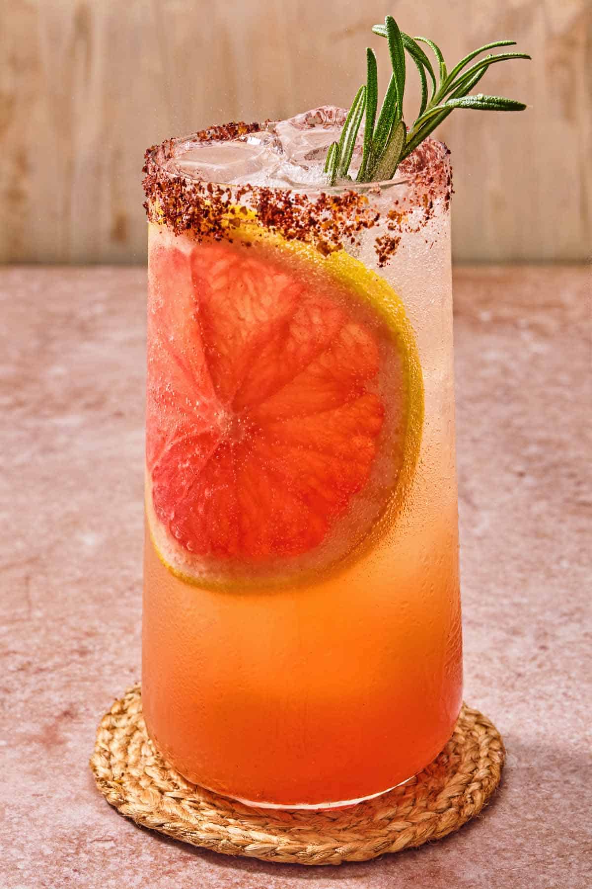 A glass of the rosemary grapefruit tonic with a grapefruit wheel, a sprig of rosemary and a sumac rim on a coaster.