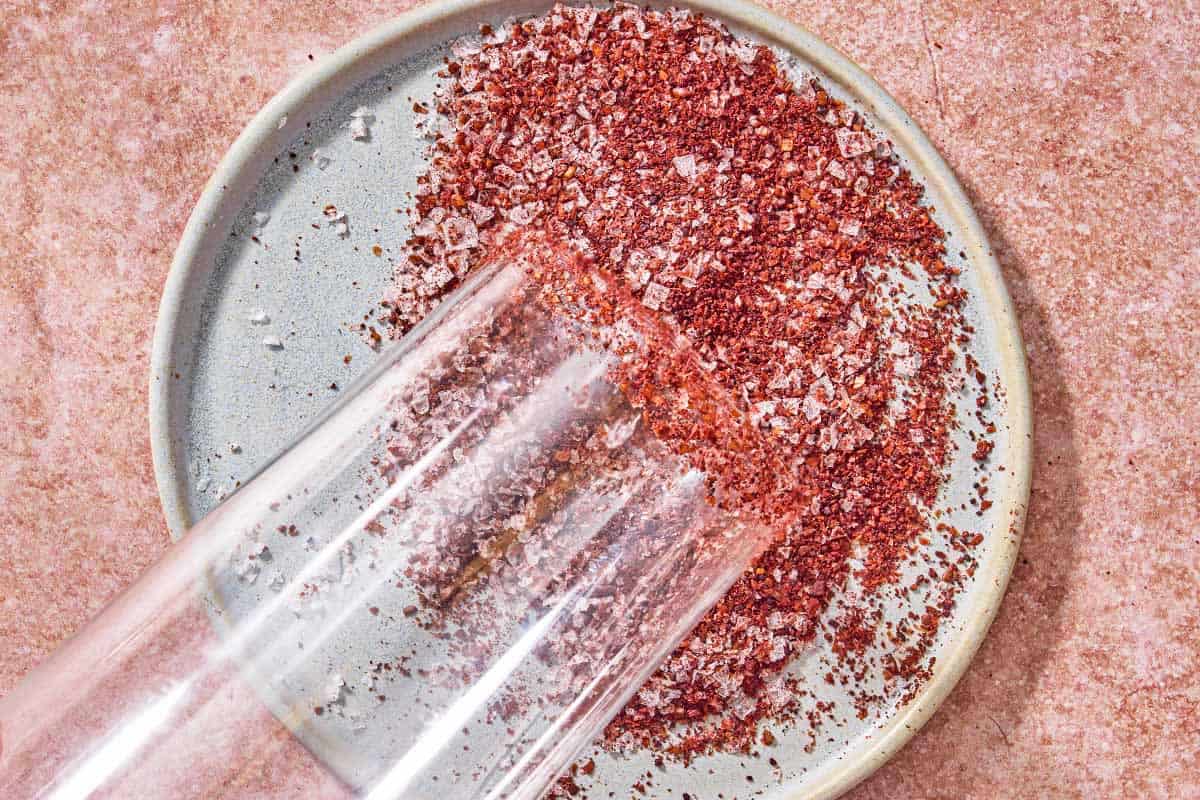 The rim of a glass being dipped in a mixture of sumac and sea salt on a small plate.