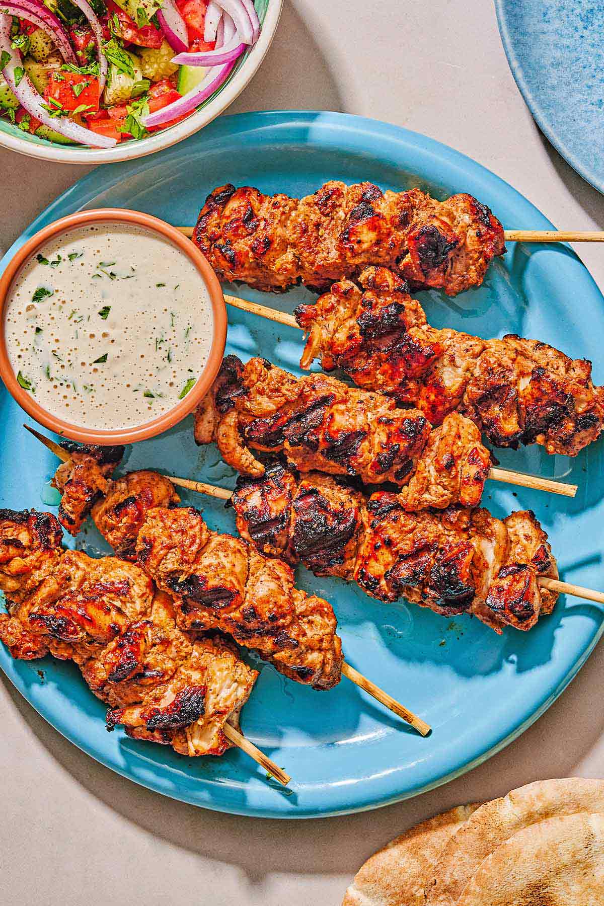 Several shish tawook skewers on a platter with a bowl of tahini sauce. Next to this is a bowl of salad and a stack of pita bread.