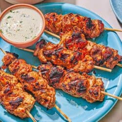 Several shish tawook skewers on a platter with a bowl of tahini sauce. Next to this is a bowl of salad and a stack of pita bread.