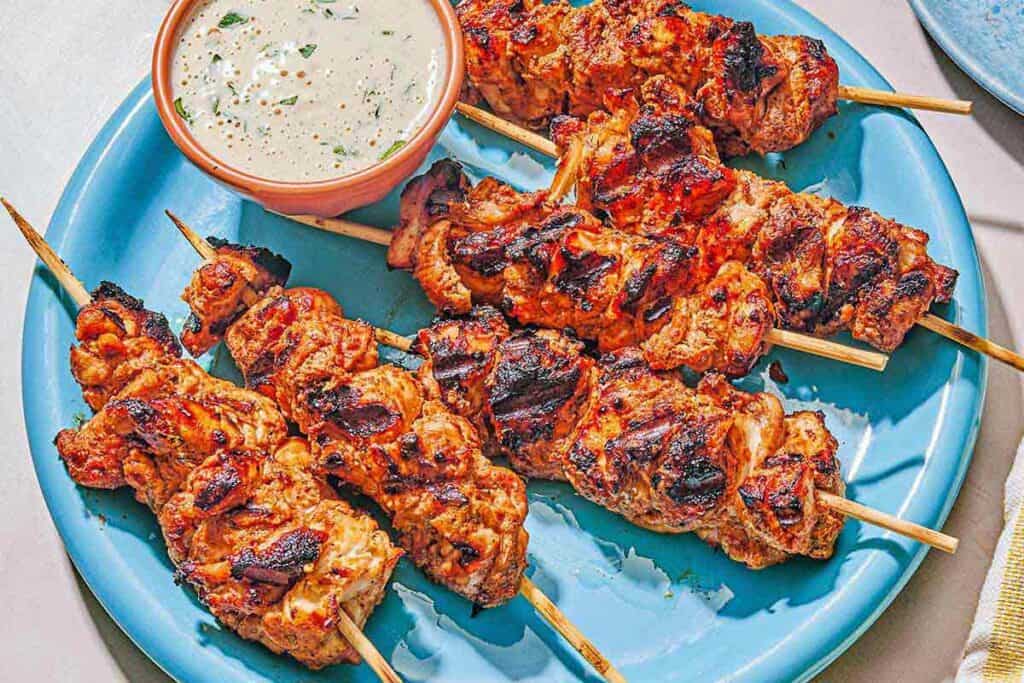 Several shish tawook skewers on a platter with a bowl of tahini sauce.
