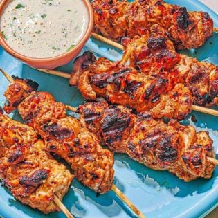 Several shish tawook skewers on a platter with a bowl of tahini sauce.