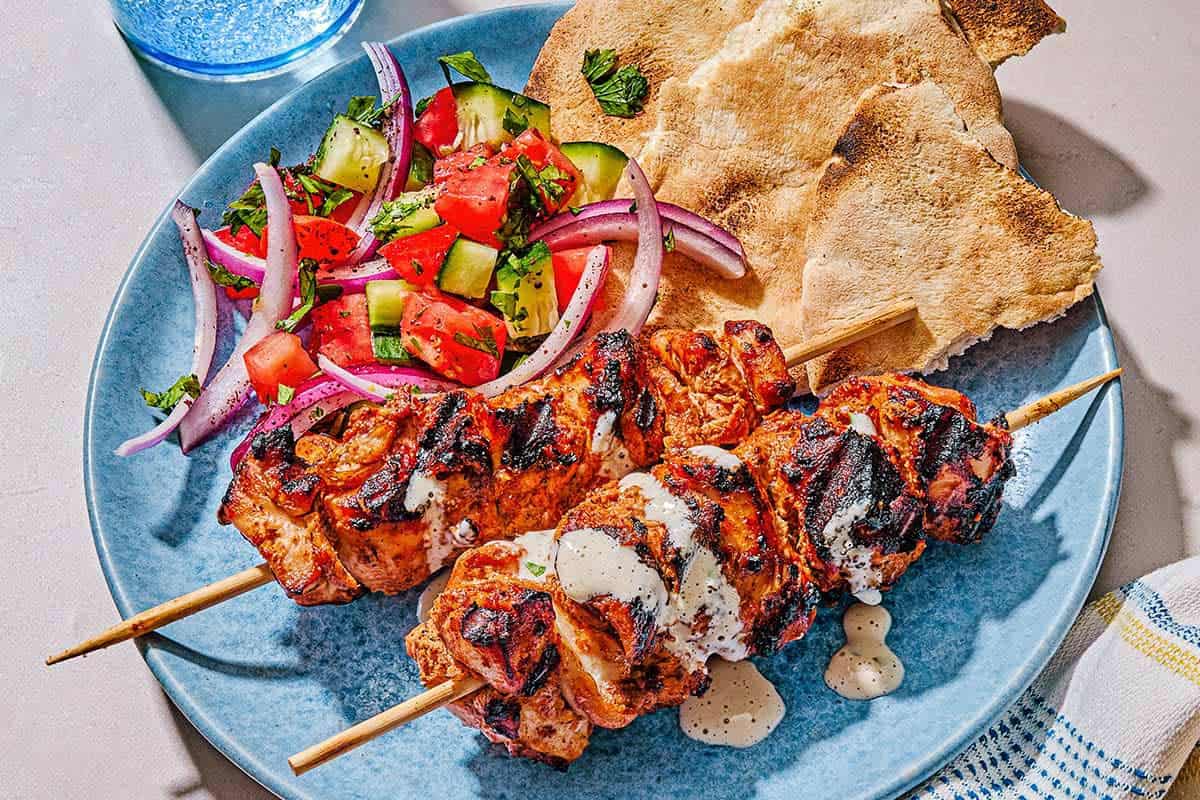 Two shish tawook skewers on a plate with a salad and pieces of pita. Next to this is a kitchen towel and a glass of sparkling water.