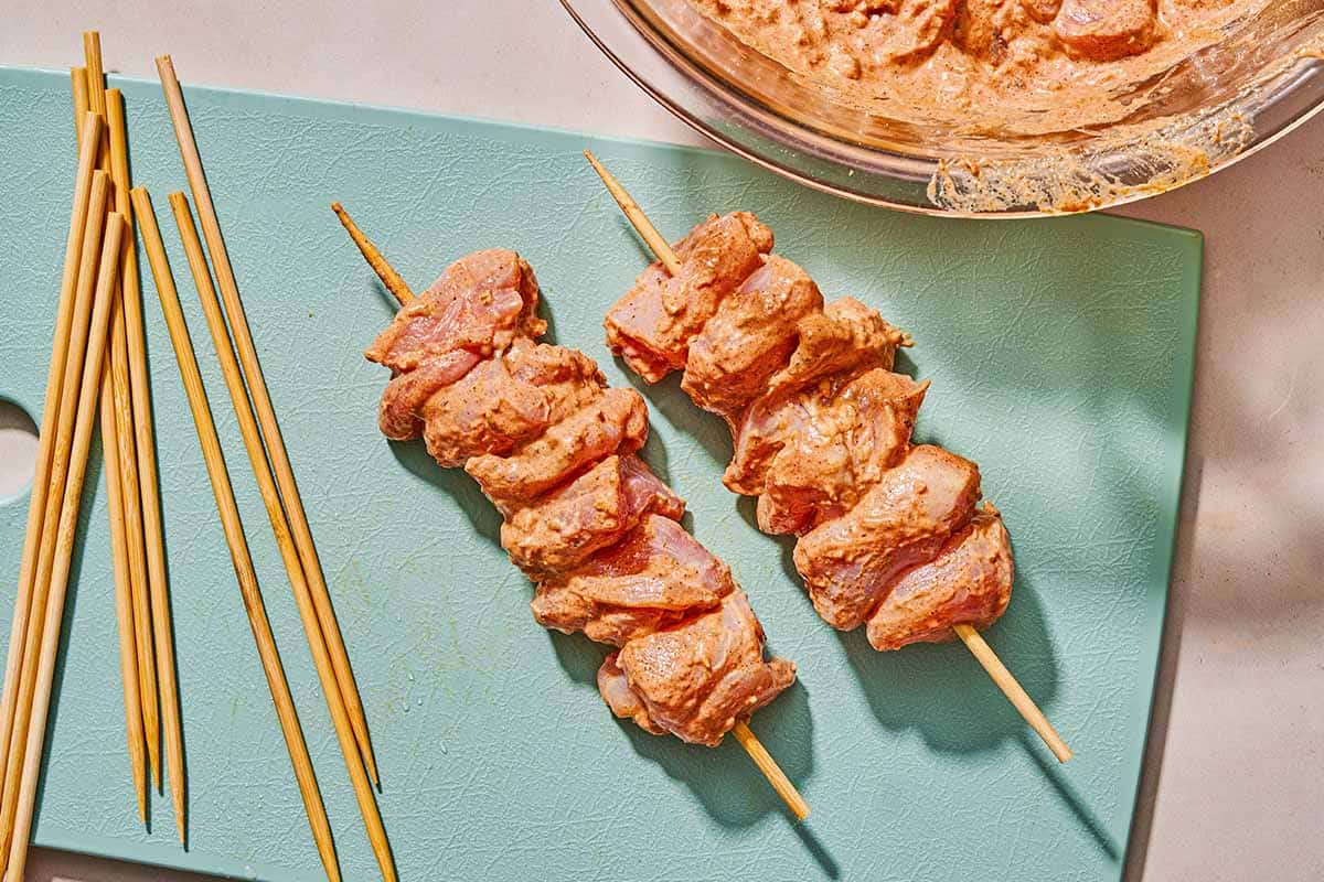 Two uncooked shish tawook skewers on a cutting board with several skewers. Next to this is the bowl of chicken thighs in marinade.