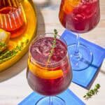 Two tart cherry juice mocktails garnished with lemon twists and sprigs of fresh thyme on coasters. Next to this is a tray with the ingredients for the mocktails.