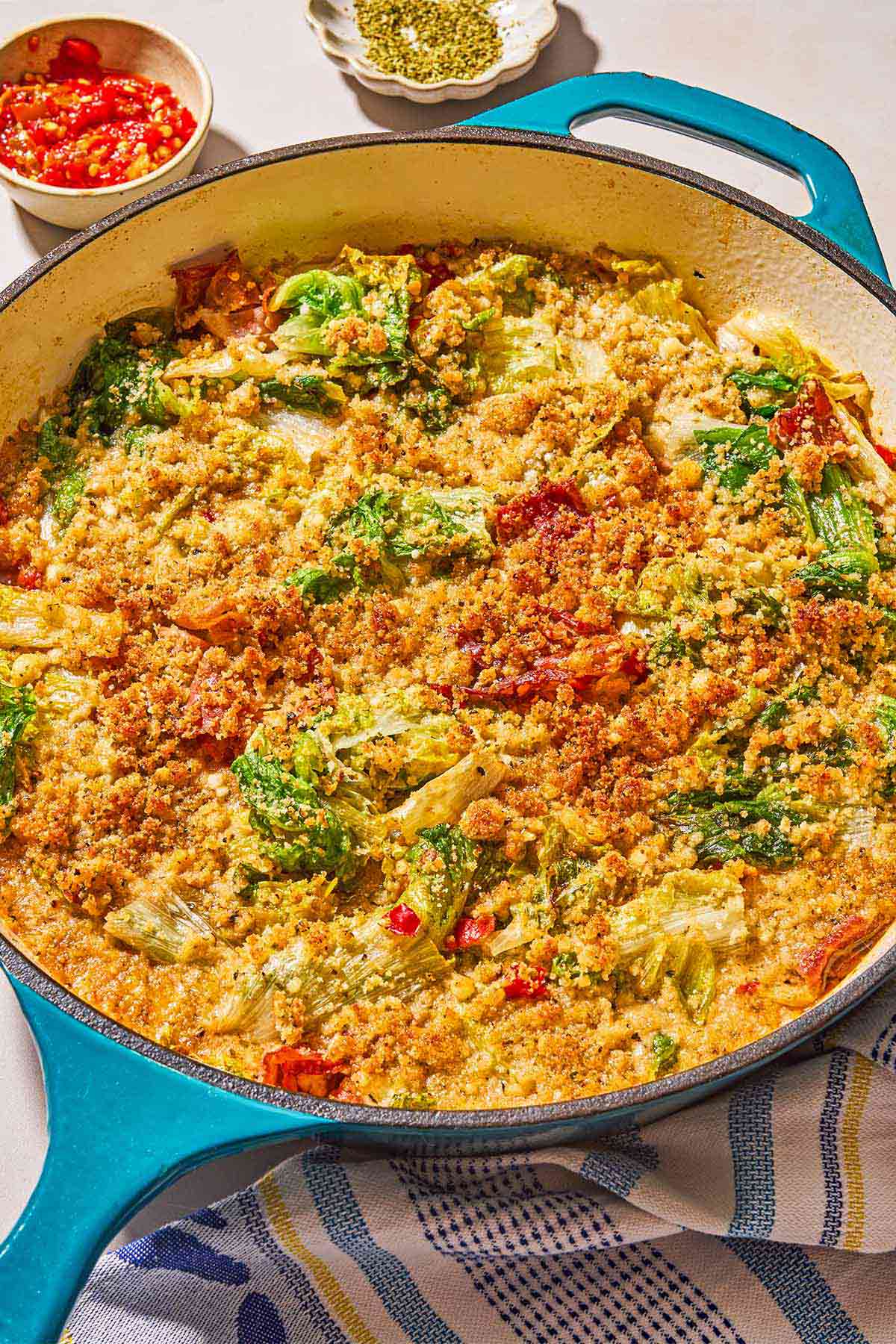 Utica greens in a skillet next to a kitchen towel, a bowl of dried oregano, and a bowl of pickled cherry peppers.