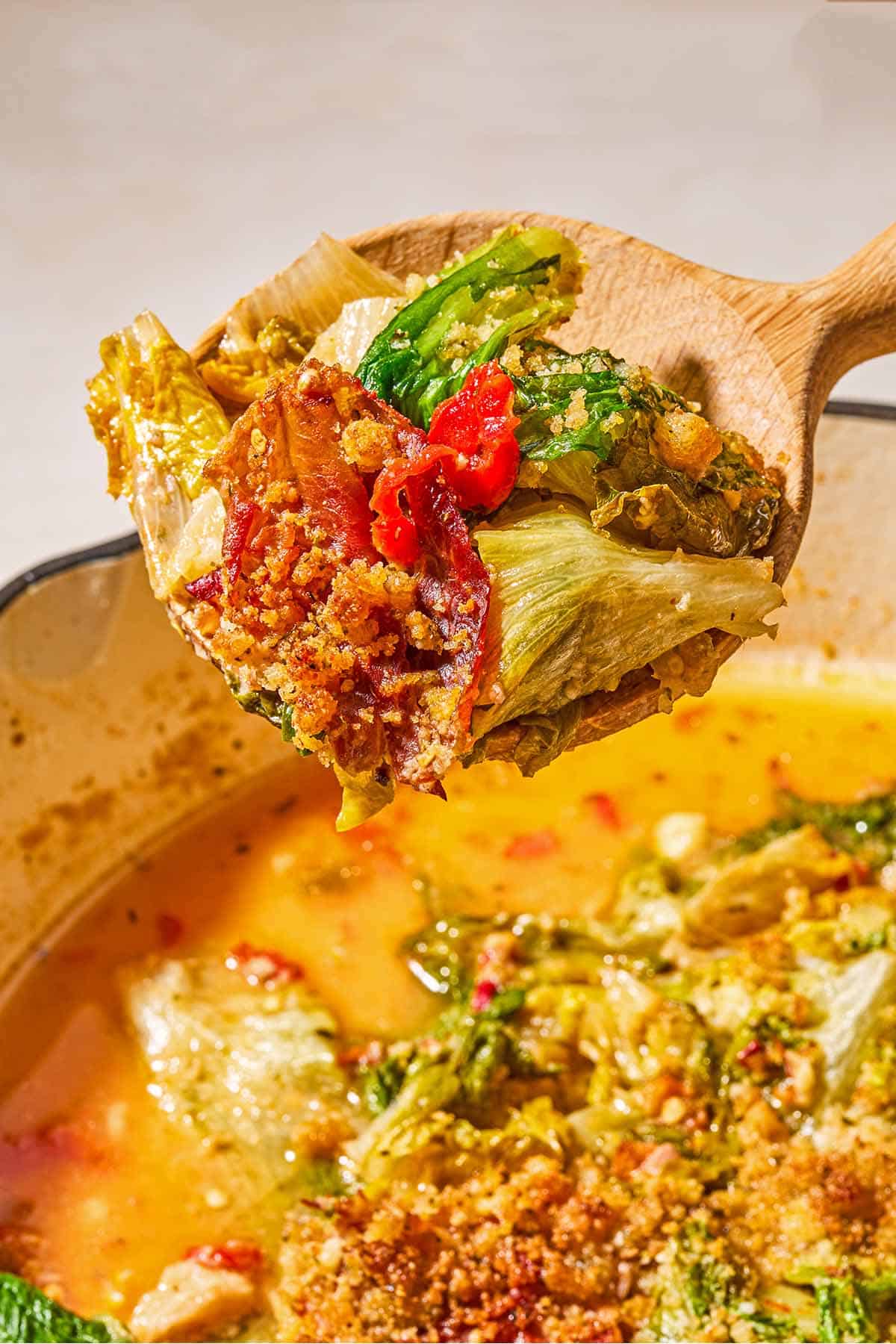 A close up of a wooden spoon with a bit of the utica greens being held over a skillet with the rest of the recipe.