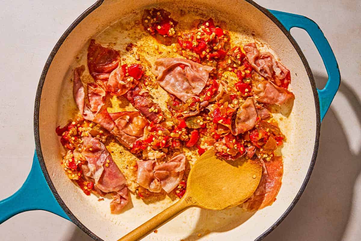 The chopped prosciutto, pickled cherry peppers and garlic being sautéed in a skillet with a wooden spoon.