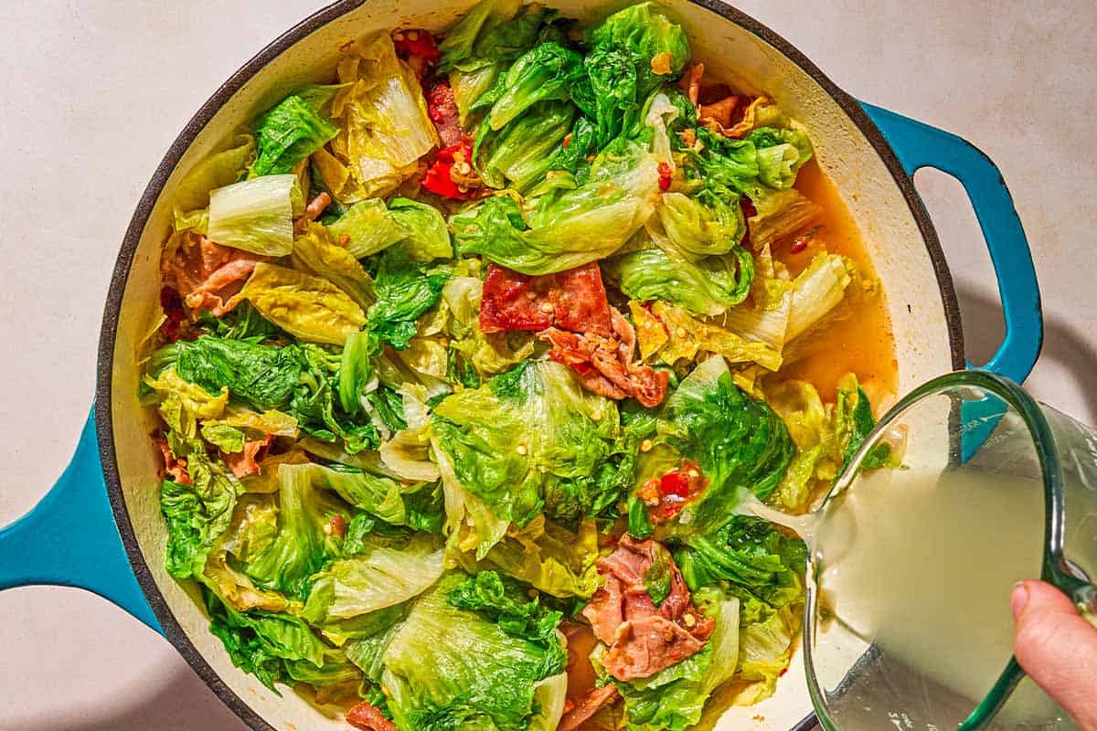 Chicken broth being poured over the aromatics and greens for the utica green recipe in a skillet.