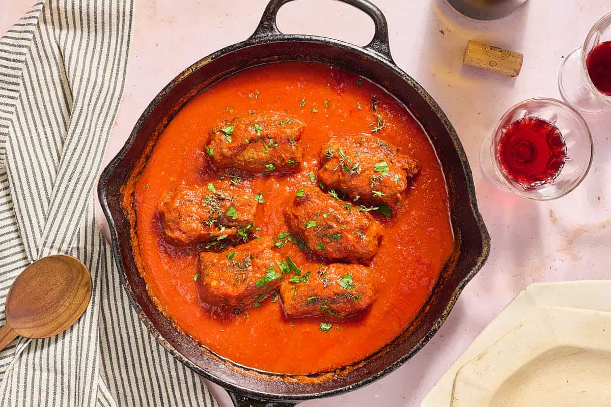 The braciole braised in marinara sauce in a cast iron skillet. Next to this is a cloth napkin, a wooden spoon, a stack of 2 plates, a glass of wine and a wine cork.